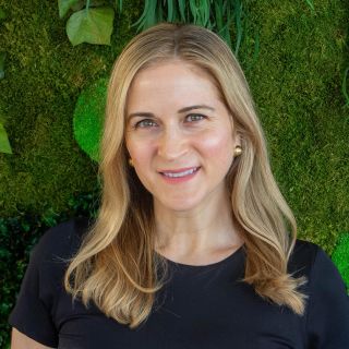 Photograph of smiling woman with blonde hair, black top, gold earrings against green plant wall