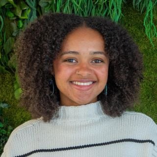 Photograph: smiling young woman with curly hair, nose ring, striped sweater, green plant wall