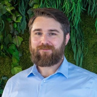 Photograph: smiling bearded man in light-blue shirt against lush green plant wall