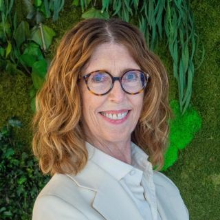 Smiling woman with glasses in beige blazer against lush green plant wall (photo)