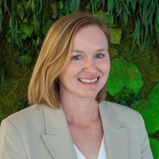 Photograph of smiling woman with blonde bob in beige blazer against green living wall
