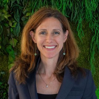 Photograph: smiling woman in navy blazer, pendant necklace, green plant background