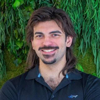 Photograph of smiling man with mullet, mustache and goatee, black polo against green foliage