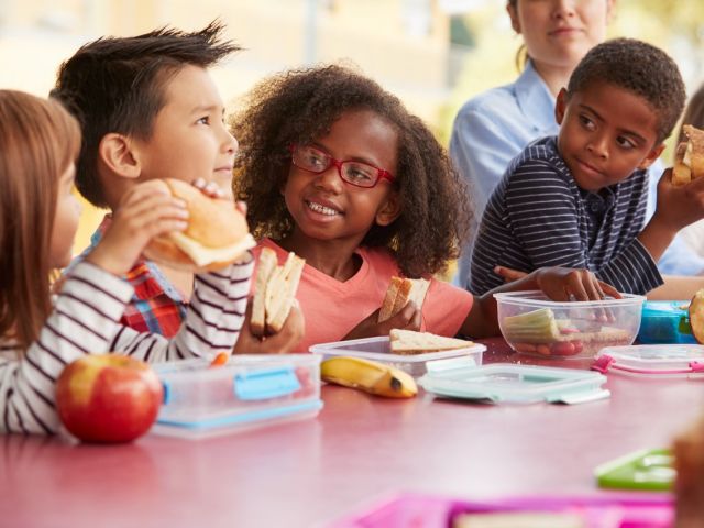 Photo: diverse elementary children at lunch table smiling and eating sandwiches.