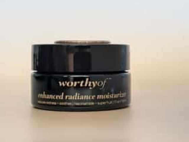 Photograph of a small black jar labeled 'enhanced radiance moisturizer' on beige background.