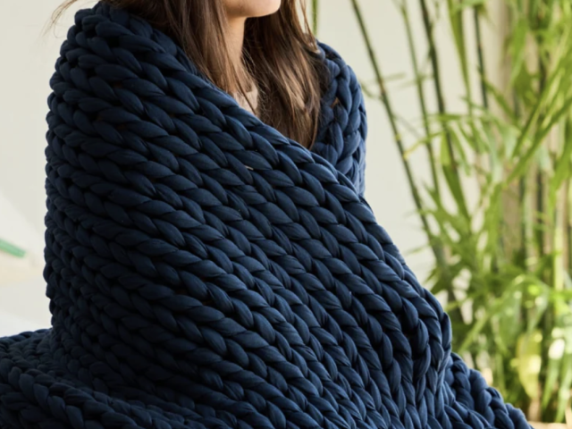 Photograph of woman wrapped in chunky navy blue knit blanket with bamboo in background