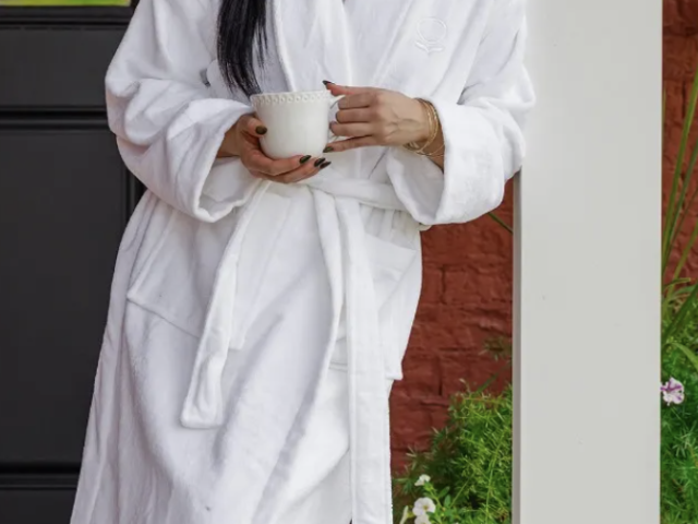 Photograph: woman in white bathrobe holding a mug on a porch beside potted flowers.