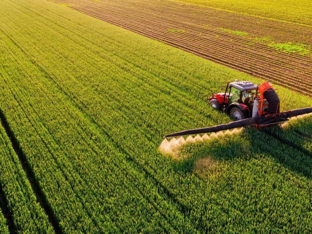 Aerial photo of a red tractor spraying a vast green crop field at golden hour.