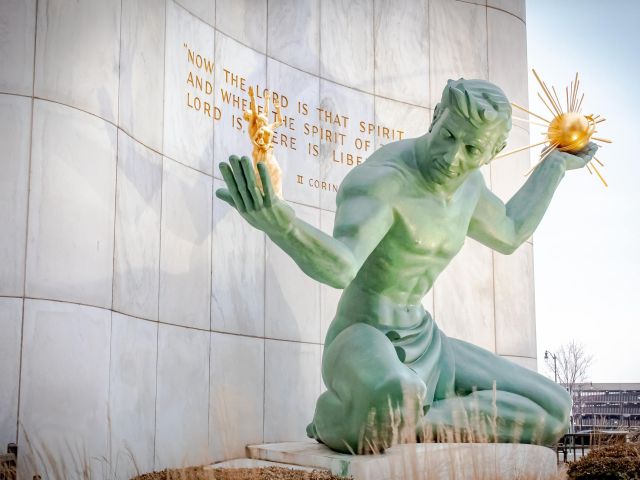 Seated green bronze statue holding golden sun and family figure against marble wall