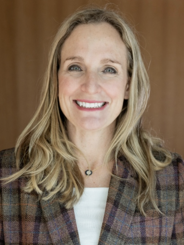 Photograph of smiling blonde woman in plaid blazer against wooden backdrop