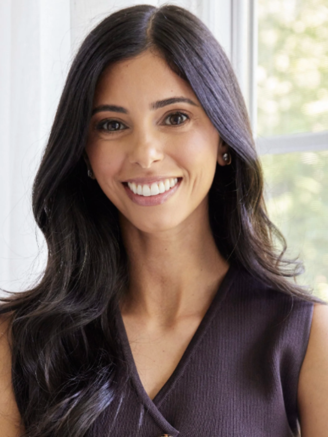 Headshot photograph: smiling woman with long dark hair by a window, wearing sleeveless dark top