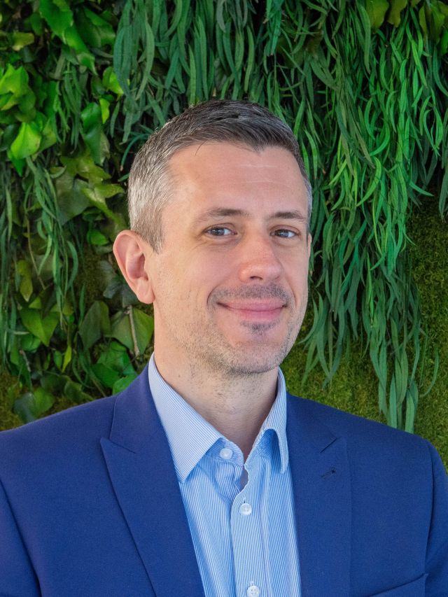 Photograph of a smiling man in blue suit before a green plant wall