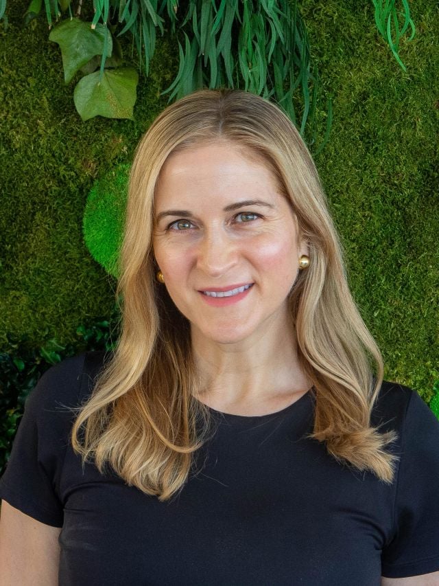 Photograph of smiling woman with blonde hair, black top, gold earrings against green plant wall