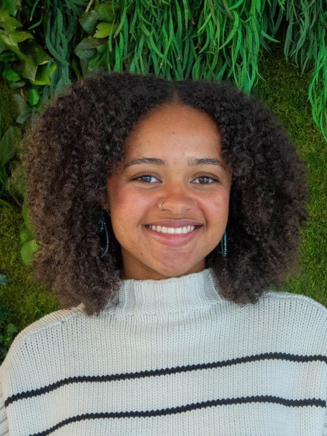 Photograph: smiling young woman with curly hair, nose ring, striped sweater, green plant wall