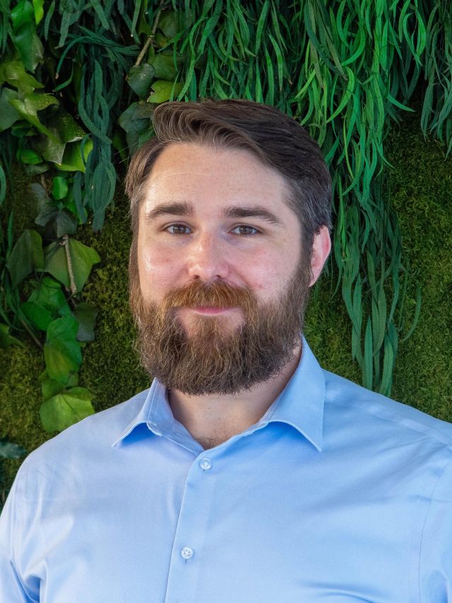 Photograph: smiling bearded man in light-blue shirt against lush green plant wall