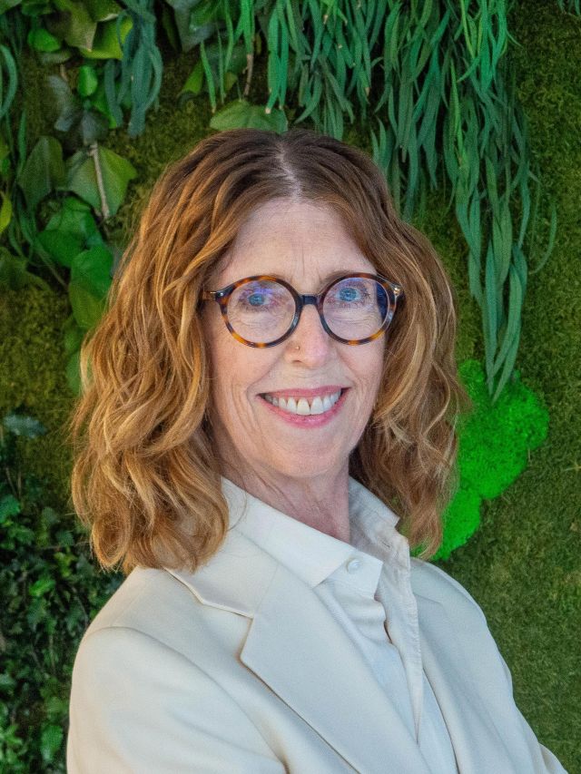 Smiling woman with glasses in beige blazer against lush green plant wall (photo)