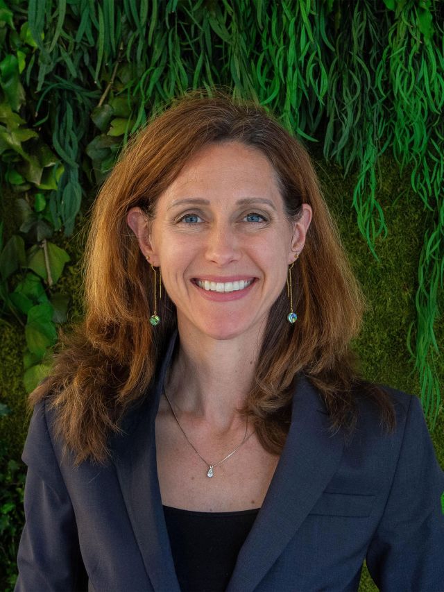 Photograph: smiling woman in navy blazer, pendant necklace, green plant background