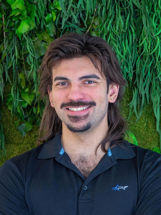 Photograph of smiling man with mullet, mustache and goatee, black polo against green foliage