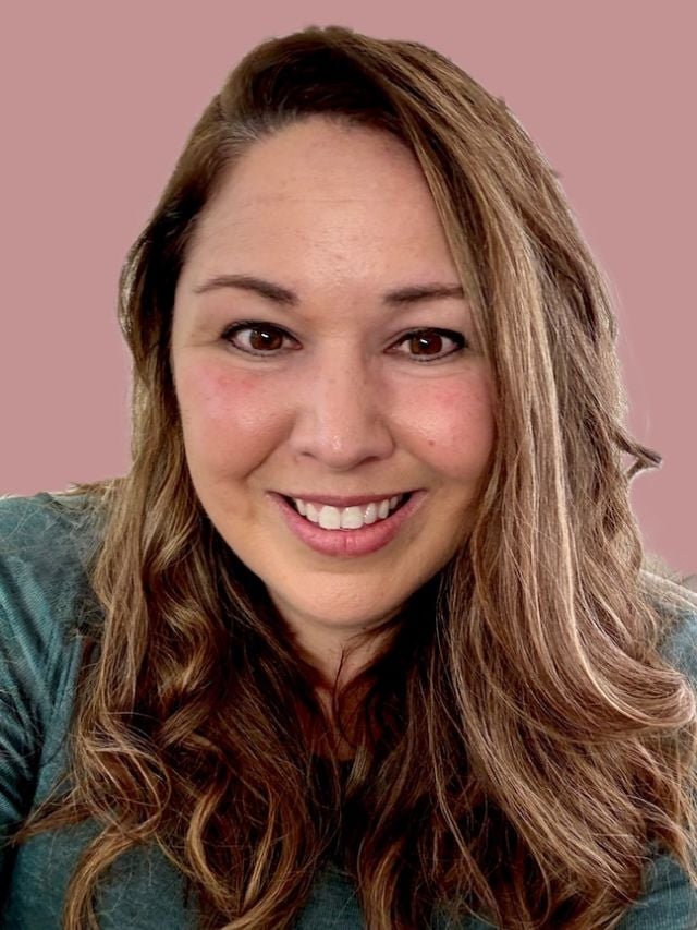 Photograph of smiling woman with long wavy brown hair, green top, pink background