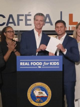 Photograph: smiling officials at cafeteria podium holding document, LAUSD logo behind