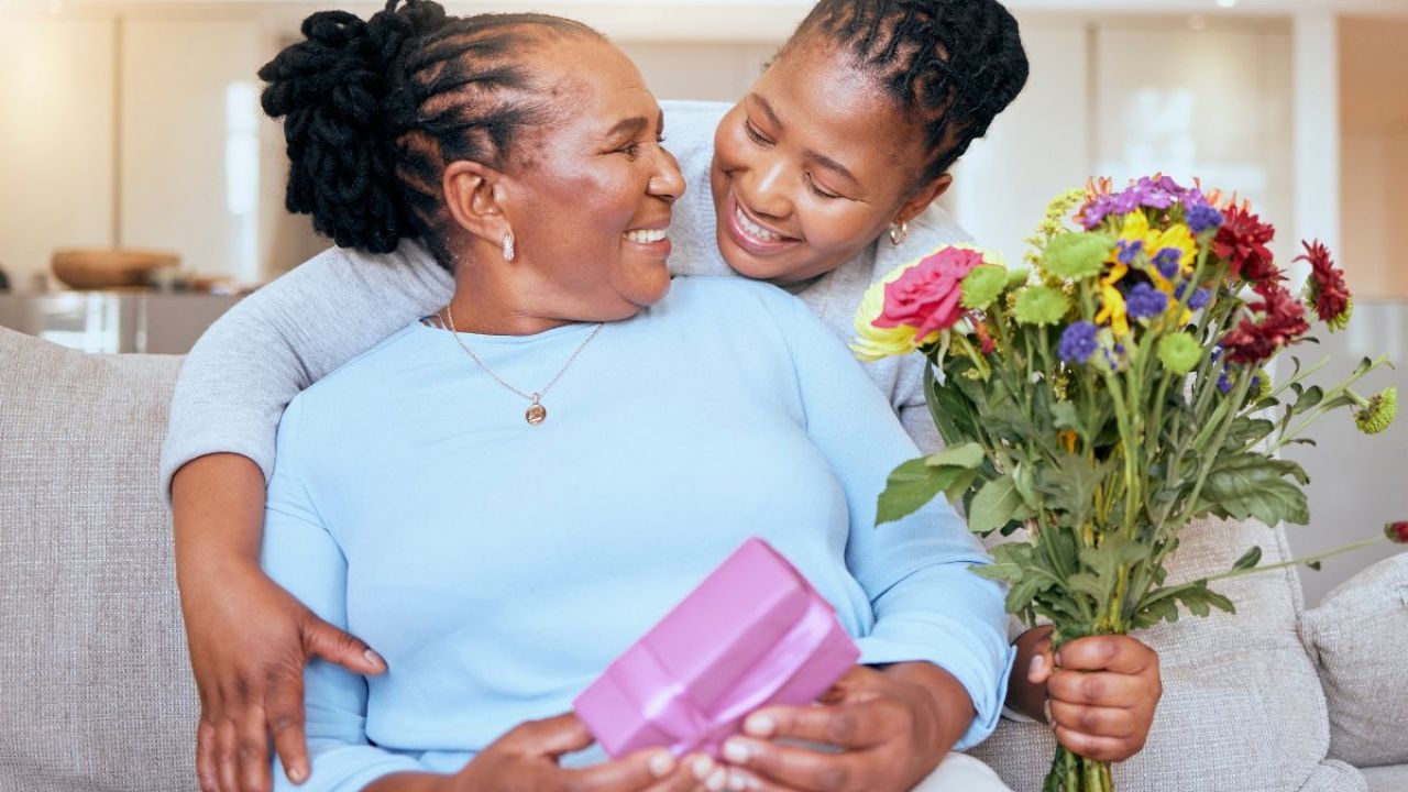 Photograph of mother and daughter smiling on couch; daughter with flowers, mother with purple gift