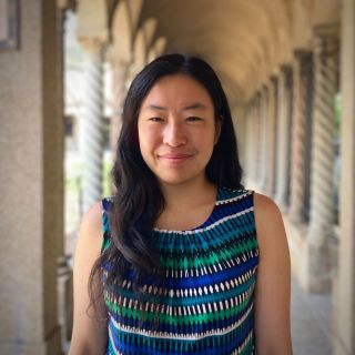 A young woman with long dark hair smiles, wearing a colorful patterned top, in a corridor.