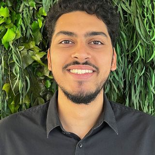 Smiling young man with curly black hair in front of a lush green wall.