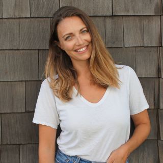 Woman with long, wavy hair and a bright smile, standing against a wooden wall.