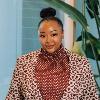 A woman with a bun hairstyle wears a patterned blazer over a maroon turtleneck, smiling.