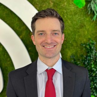 Smiling man in a suit and red tie, standing against a green foliage backdrop.
