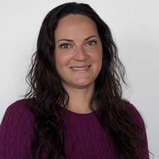 Smiling woman with long, dark, wavy hair wearing a purple sweater against a white background.