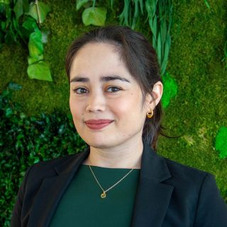 A woman in a black blazer smiles against a green, plant-filled backdrop.