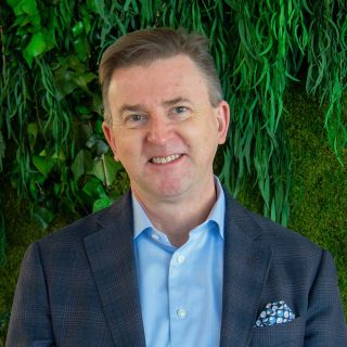 A smiling man in a blazer stands against a backdrop of green foliage.