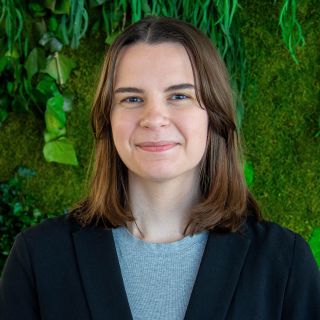 A young woman with shoulder-length brown hair smiles, dressed in a black blazer against a green wall.