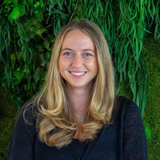 Smiling woman with long, wavy hair in front of a lush green plant backdrop.