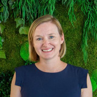 Smiling woman with short blonde hair, wearing a navy blue top, against a green botanical background.
