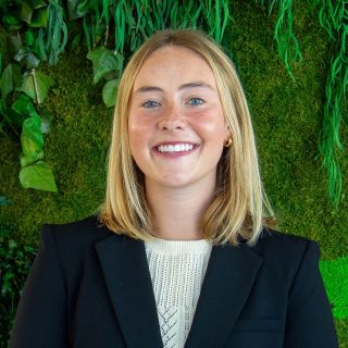Smiling woman with blonde hair in a black blazer, posing against a green plant background.