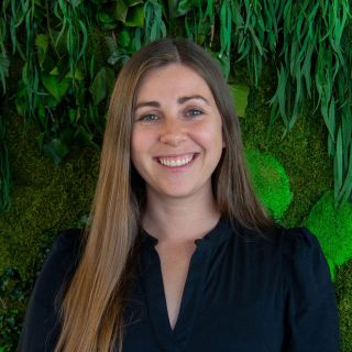 Young woman with long brown hair smiling in front of a green plant backdrop.