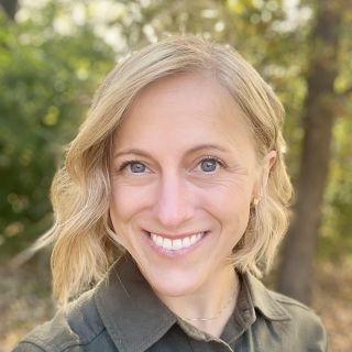 Smiling woman with wavy blonde hair standing outdoors among green trees.