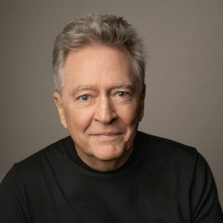Headshot photograph of man with gray hair, black sweater, neutral background