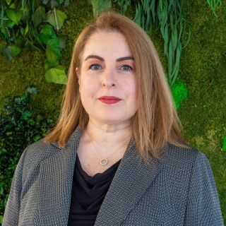 Photograph: woman with shoulder-length light brown hair, gray blazer, against green plant wall