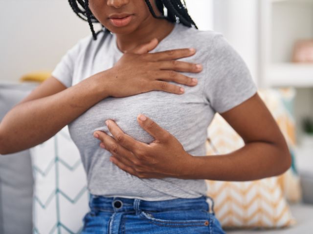 A woman sitting on a couch holds her chest, expressing discomfort or concern.