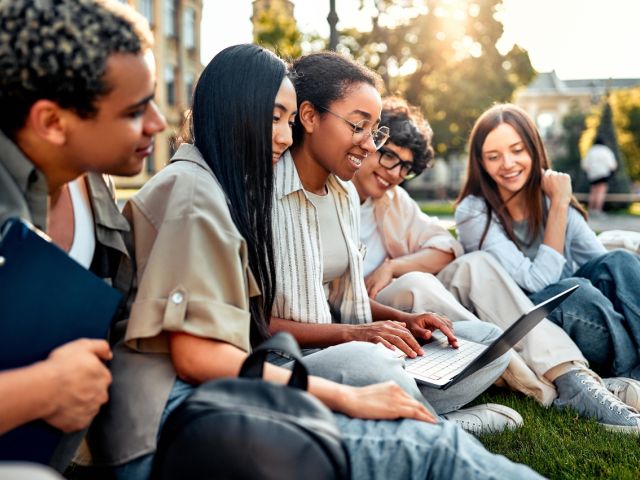 A diverse group of five people collaborates outdoors, smiling and working on a laptop.