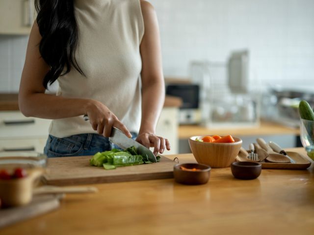 A person chops vegetables on a wooden kitchen counter, surrounded by various bowls of food.