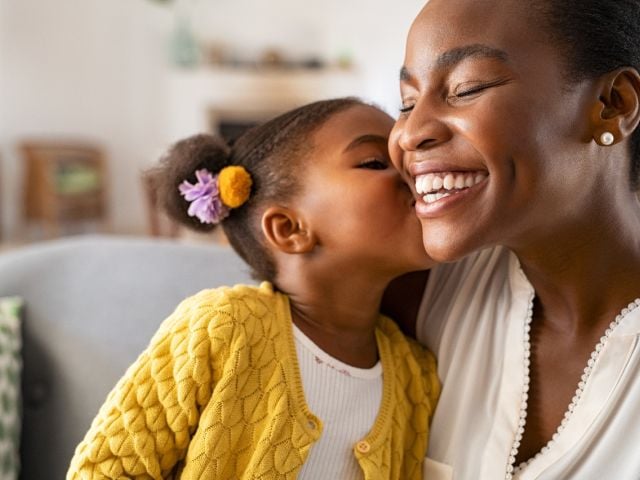 A mother smiles joyfully as her daughter kisses her cheek, both appearing cheerful.