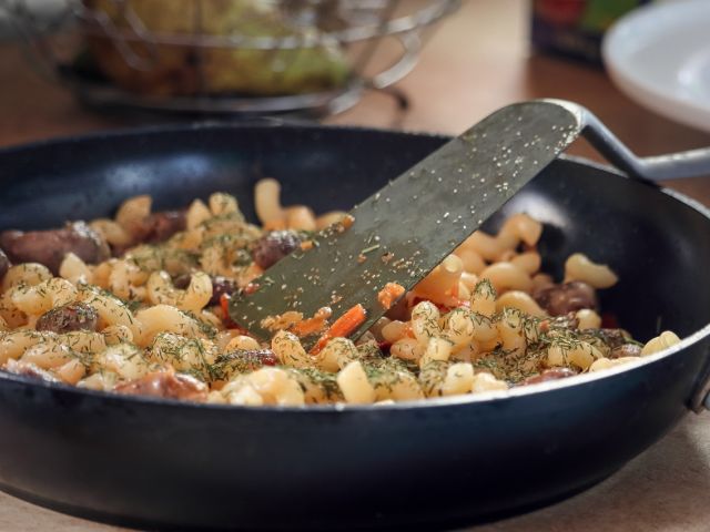 A skillet filled with pasta, vegetables, and herbs being stirred with a spatula.