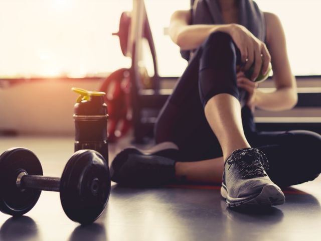 A person sits on a gym floor beside a dumbbell and a water bottle, resting.
