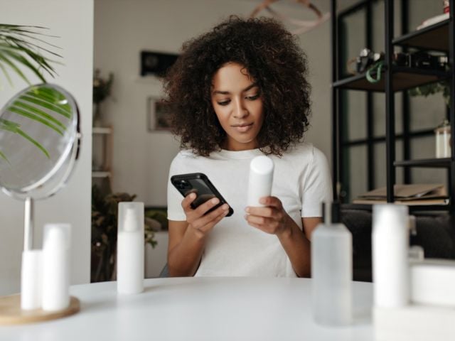 Woman with curly hair checking her phone while holding a skincare product.