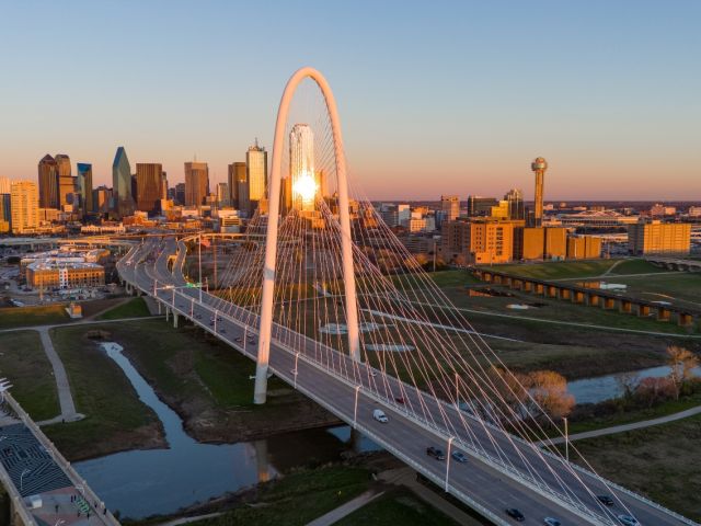Dallas skyline at sunset with a modern suspension bridge in the foreground.