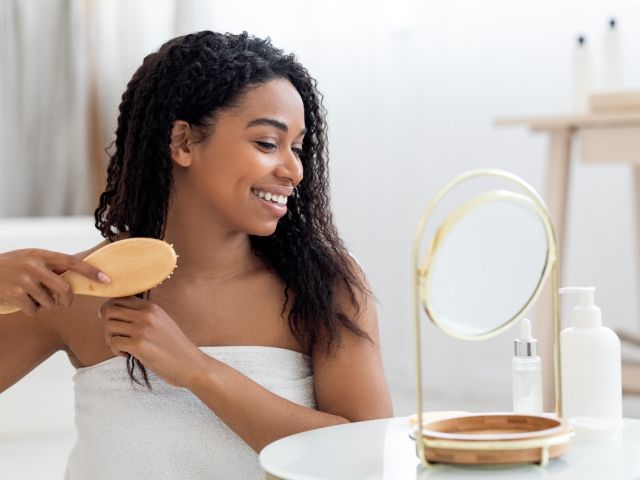 A young woman brushes her curly hair while smiling in front of a mirror.